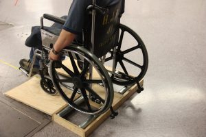 A manual wheelchair stands on a platform with two pairs of rollers